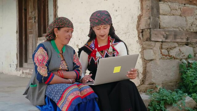 Rural Asian Indian daughter in traditional Hindu costume teaching ole woman or mother how to use a laptop in a remote or village area. Concept of the countryside, village online education learning.
