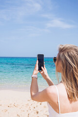 A young woman seen from back holding a smartphone with black display on blue sky and sea background. Working from home, outdoor office concept. Digital detox.