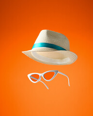 Straw hat and elegant sunglasses floating in air on bright orange background. Concept of fashion clothing accessories for beach holidays, vacations and travel.