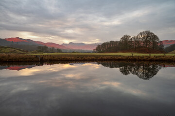 Fototapeta premium Beautiful morning light on Cumbrian mountains reflecting in calm river on a peaceful morning at The River Brathay in The Lake District, UK.