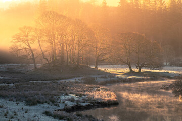 Breathtaking golden light illuminating frosty Winter landscape with misty river near Elterwater in...