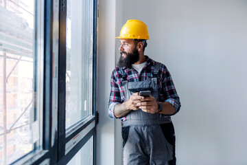 A happy, bearded worker with a helmet on his head, is standing next to a window and using his phone...