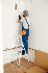 Electrician, handyman standing on a ladder while working on installing socket on wall at home
