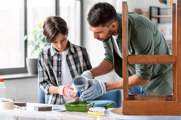 repair, diy and home improvement concept - father and son in protective gloves pouring grey color paint into tray for painting old wooden table at home