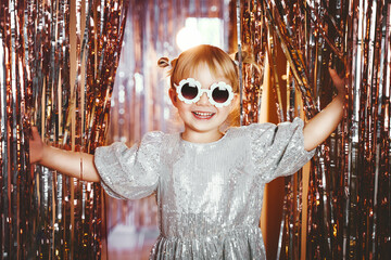 Happy little stylish girl in shiny dress having fun. Festive background with foil curtain...