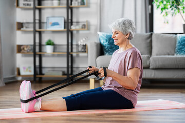 sport, fitness and healthy lifestyle concept - smiling senior woman exercising with resistance band on mat at home - obrazy, fototapety, plakaty