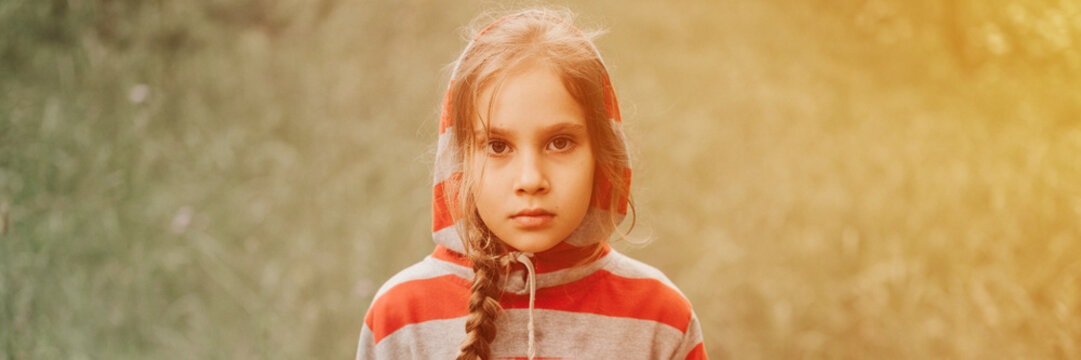 portrait face of upset or focused thoughtful scowl eight year old kid girl in hood of striped hoodie in the forest nature. prepubertal children mind psychology and mental health. banner. flare