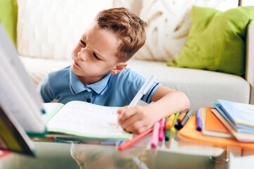 Thinking child bored, little boy writing carefully sitting at desk and doing homework