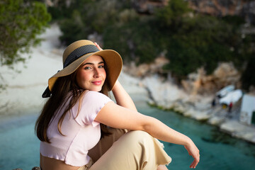 Happy traveller with a hat in front of the beach at sunset looking to the camera. Handsome woman portrait on a cliff.