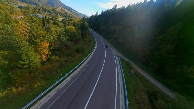 Travel drone flight. FPV aerial shot of speed biker riding fast motorcycle bike on country highway. Sunny sky. Picturesque forests. Mountains.