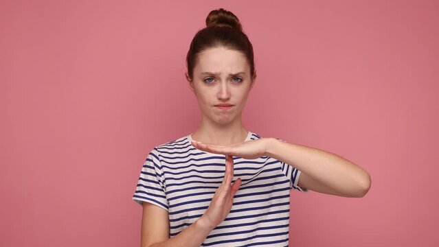 I need more time, enough limits. Displeased woman showing time out hand gesture, looking imploringly, worried about deadline, wearing striped T-shirt. Indoor studio shot isolated on pink background.