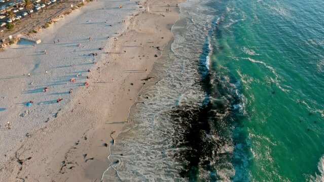 Aerial shot flying forward over Camps Bay beach while tilting up to reveal the  beachfront's full splendour and the Twelve Apostle mountain range in the background during sunset