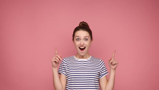 Wow, look above at advertisement. Pleasantly surprised girl pointing up and looking with shocked face, showing copy space for commercial. Indoor studio shot isolated on pink background.