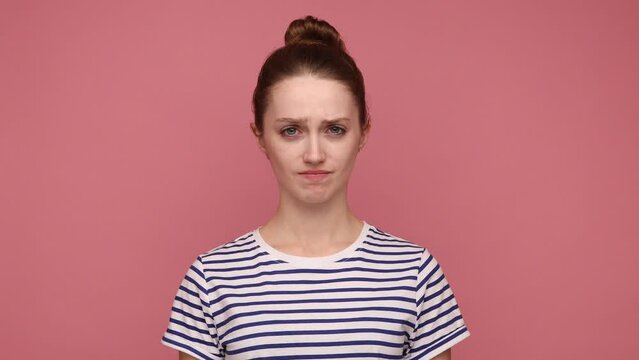 Serious attractive woman with hair bun nodding no, expressing disagreement, looking at camera with rejection, wearing striped T-shirt. Indoor studio shot isolated on pink background.