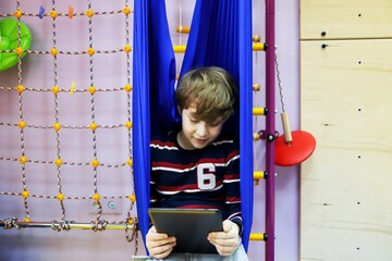 child boy watching cartoons on a tablet while sitting in a hammock at a children's center for the development of special children