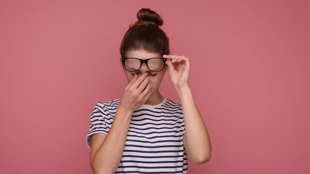 Attractive exhausted woman with hair bun takes off glasses, feels eyes pain, being tired from long hours working, wearing striped T-shirt. Indoor studio shot isolated on pink background.
