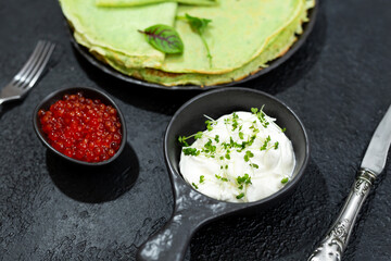 Classic thin pancakes made of green spinach with sour cream and red caviar in a black plate. Top view