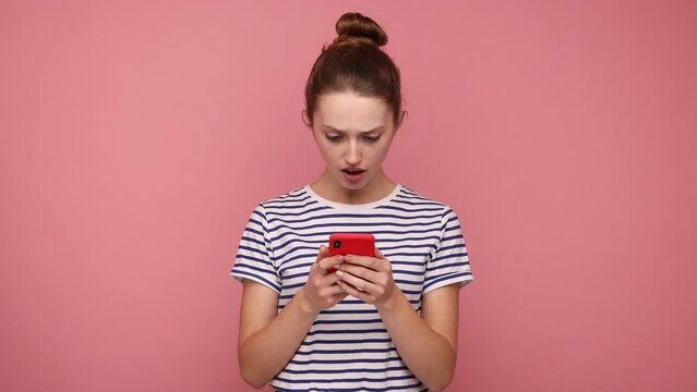 Shocked astonished woman with hair bun reading post on social network using cell phone, chatting, looking surprised, wearing striped T-shirt. Indoor studio shot isolated on pink background.