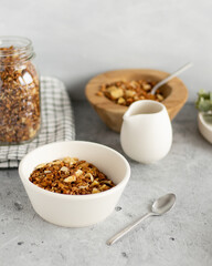 Granola in a white bowl on grey background nearby jar of granola, pitcher and wooden bowl with granola