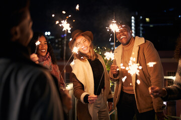 Young adults laughing at a rooftop party. Multi-cultural group of friends lighting sparklers with city scape background