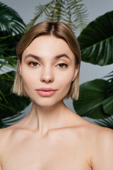 Fototapeta premium pretty young woman with bare shoulders near green tropical leaves on grey