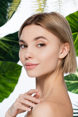 blonde woman with perfect skin looking at camera near palm leaves on white