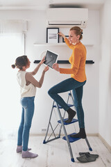 Mother and daughter on the ladder hanging pictures and photos on the shelf and wall at home.