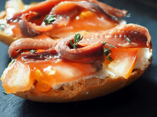 Homemade sandwiches with cheese, tomato slices and salted anchovy filets on a black stone board and on a black wooden background. Close-up