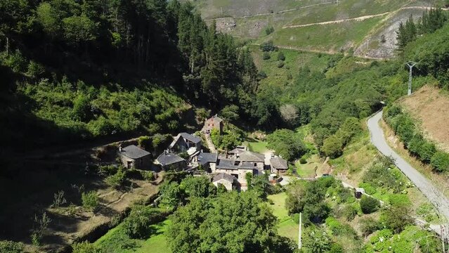 Aerial shot of drone getting very close to a little town on the valley of the green mountains of Asturias, Spain. Thic forest covering mountain surface under a clear blue sky in pure daylight.