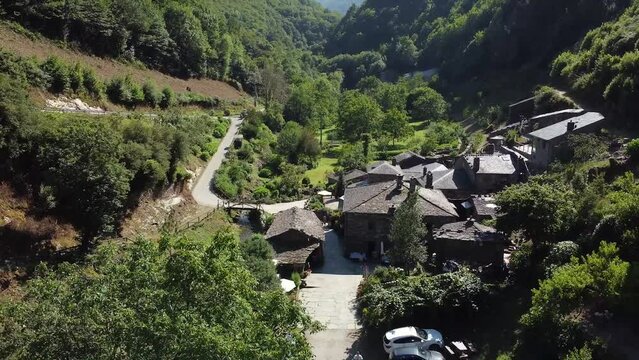Backward aerial shot from a little town (backward) on the valley of the green mountains of Asturias, Spain. Thic forest covering mountain surface under a clear blue sky in pure daylight.