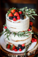 Delicious festive naked layer cake with buttercream Decorated with fresh blueberries, raspberries, strawberries, and greenery Two-tiered white wedding cake with fresh berries 