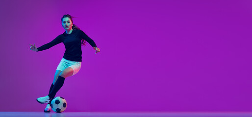 Fototapeta na wymiar Professional female soccer player dribbling football ball isolated on purple studio background in neon light. Sport, action, motion, fitness
