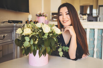 Beautiful Asian woman sits by the table in living room bright interior admires white roses flowers bouquet. Smiling young lady happy to receive a surprise gift