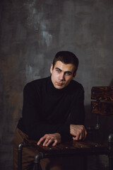 Portrait. Young man with a short hair in a black turtleneck stand near chair and looking away from the camera. High quality photo