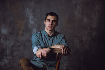 Portrait. Young man with a short hair in a turquoise shirt sit on a chair and looking in the camera. High quality photo