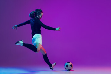 Plakat Dynamic portrait of female soccer player practicing with football ball isolated on purple studio background in neon light. Sport, action, motion, fitness