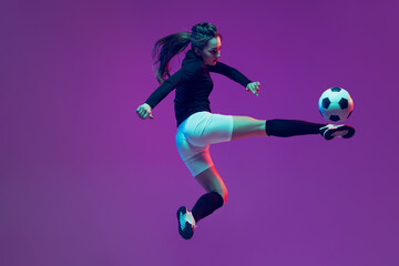 Fototapeta na wymiar In action. One sportive girl, female soccer player training with football ball isolated on purple studio background in neon light. Sport, action, motion, fitness