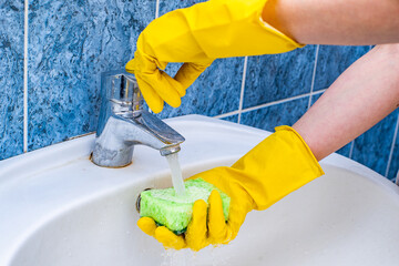 The hand of a maid or cleaning lady cleans a modern new sink in the bathroom, cleans the water tap with yellow gloves and a green sponge.