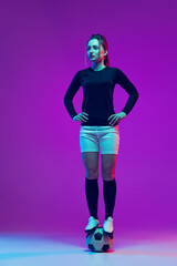 Portrait of beautiful girl, professional female soccer player posing with football ball isolated on purple studio background in neon light. Sport, team, games, fitness