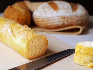 On a wooden board, slices for food are cut from a yellow wheat baguette with a bread knife. Loaves of rye bread are visible in the background.