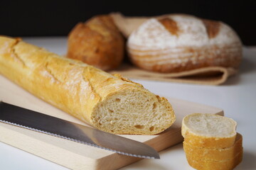 fresh baguette sliced ​​diagonally on a wooden board, a bread knife lies nearby, a little further there are two more loaves of bread