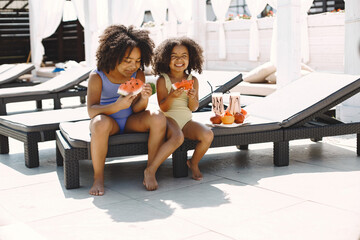 Two african american sisters sitting on a beach lounger holding a watermelon