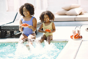 Two african american sisters sitting near the pool and holding a peace of watermelon