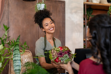 Happy black florist selling flowers to young woman - obrazy, fototapety, plakaty