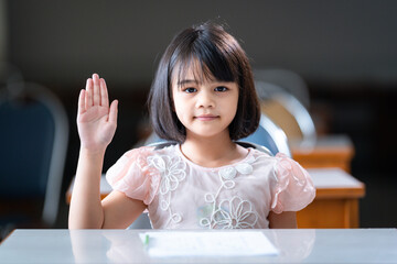 A female Asian kid student raising a hand to ask teacher question in the classroom