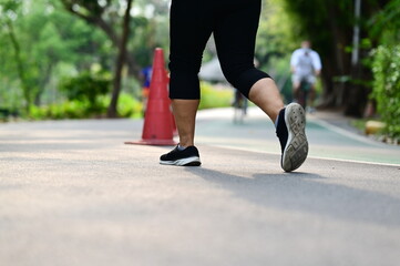 A middle-aged woman in three-quarter shorts and black sneakers jogging in a park in the fresh morning sun. Woman running slowly through green trees, orange traffic cones. (used for zoning) and cyclist