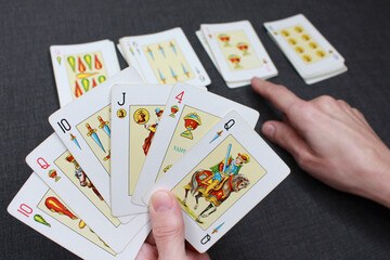A hand holding a deck of Spanish-suited cards. In the background, more cards from the deck are shown and a finger points to one of them. Colors: blacks, white and yellow.