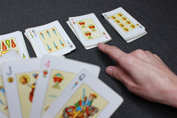 A hand holding a deck of Spanish-suited cards. In the background, more cards from the deck are shown and a finger points to one of them. Colors: blacks, white and yellow.