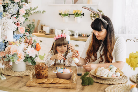 Cute little girl in bunny ears with her mother paint eggs while sitting at the table in the kitchen. Decorations for the celebration of Easter