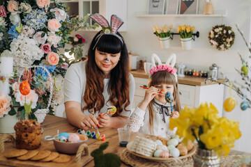 Cute little girl in bunny ears with her mother paint eggs while sitting at the table in the kitchen. Decorations for the celebration of Easter